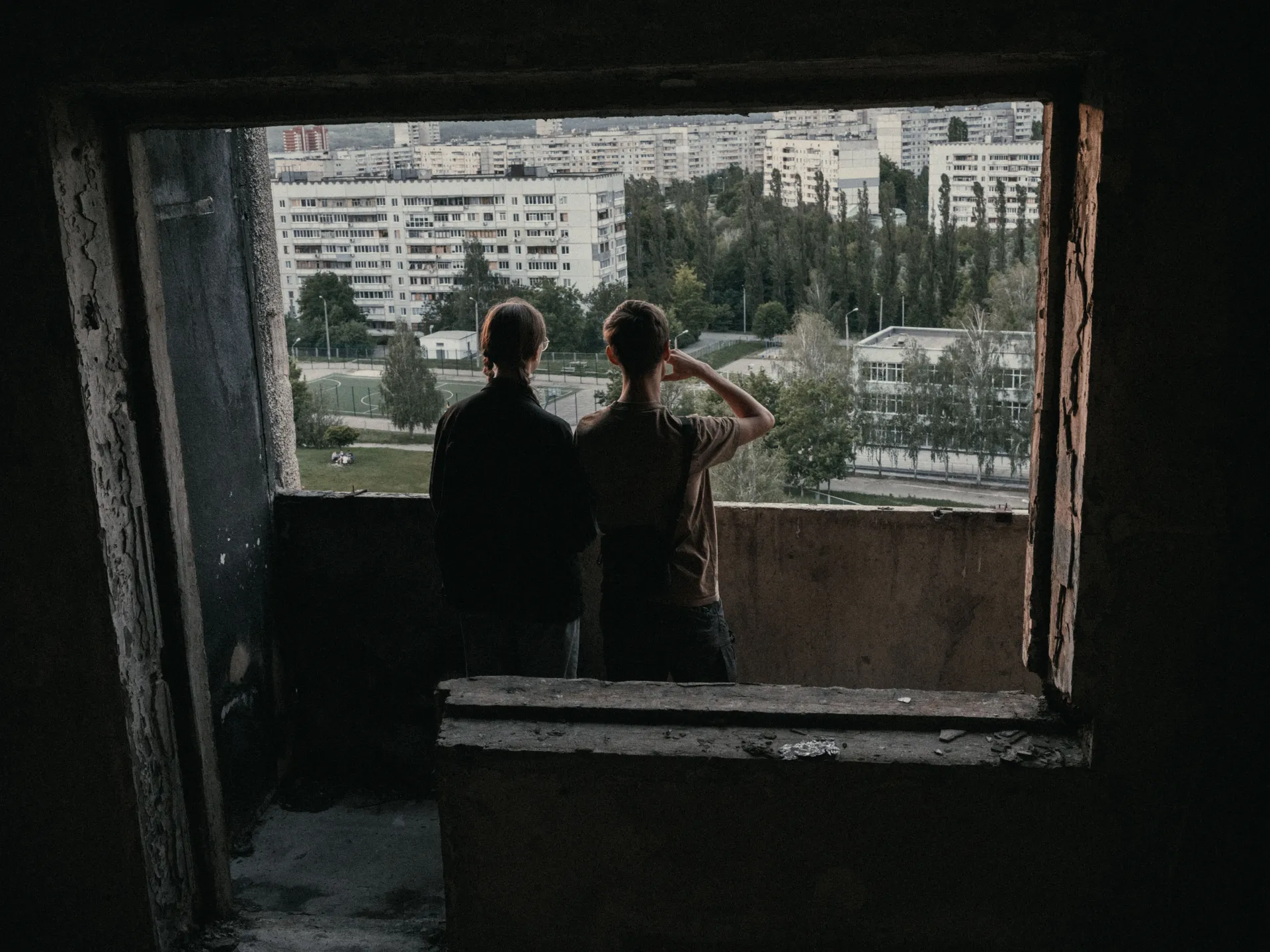 Illia, 17, and his friend Nastia talk and look out over the area from a damaged apartment building in Northern Saltivka in Kharkiv, Ukraine, on May 27, 2025. In the background is the school where Illia once studied, now reconstructed. Before Russia’s full-scale invasion, he lived in this neighborhood with his family. (George Ivanchenko / The Kyiv Independent)